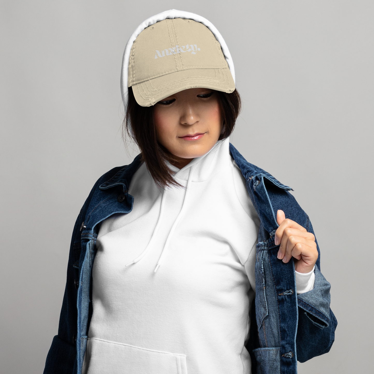 Person wearing a beige cap with text, white hoodie, and denim jacket on a gray background