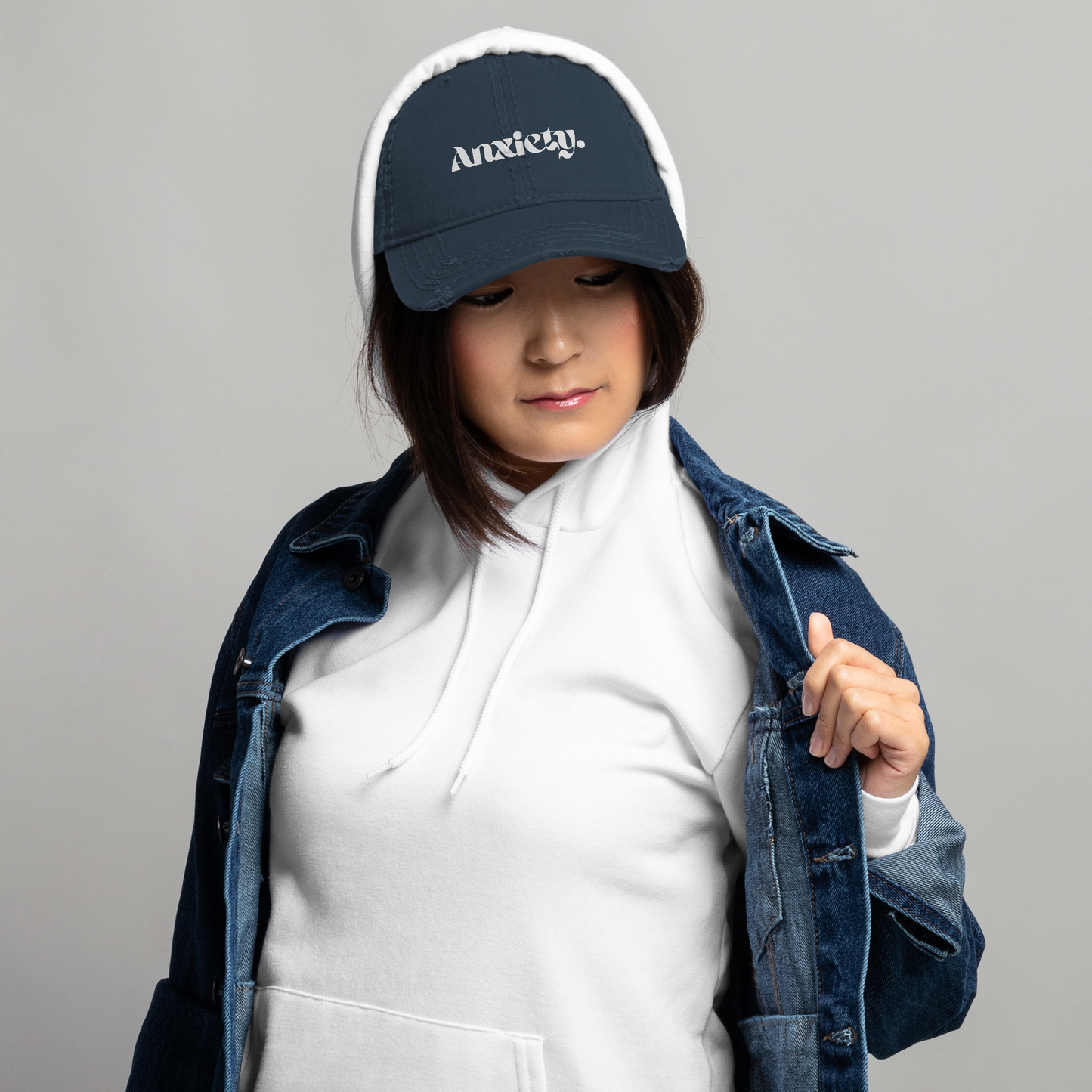 Person wearing a navy cap with 'Anxiety' text, white hoodie, and denim jacket on a gray background