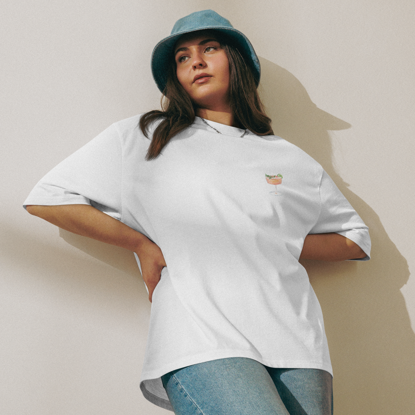 Person wearing a white t-shirt and blue jeans with a green bucket hat against a beige wall.
