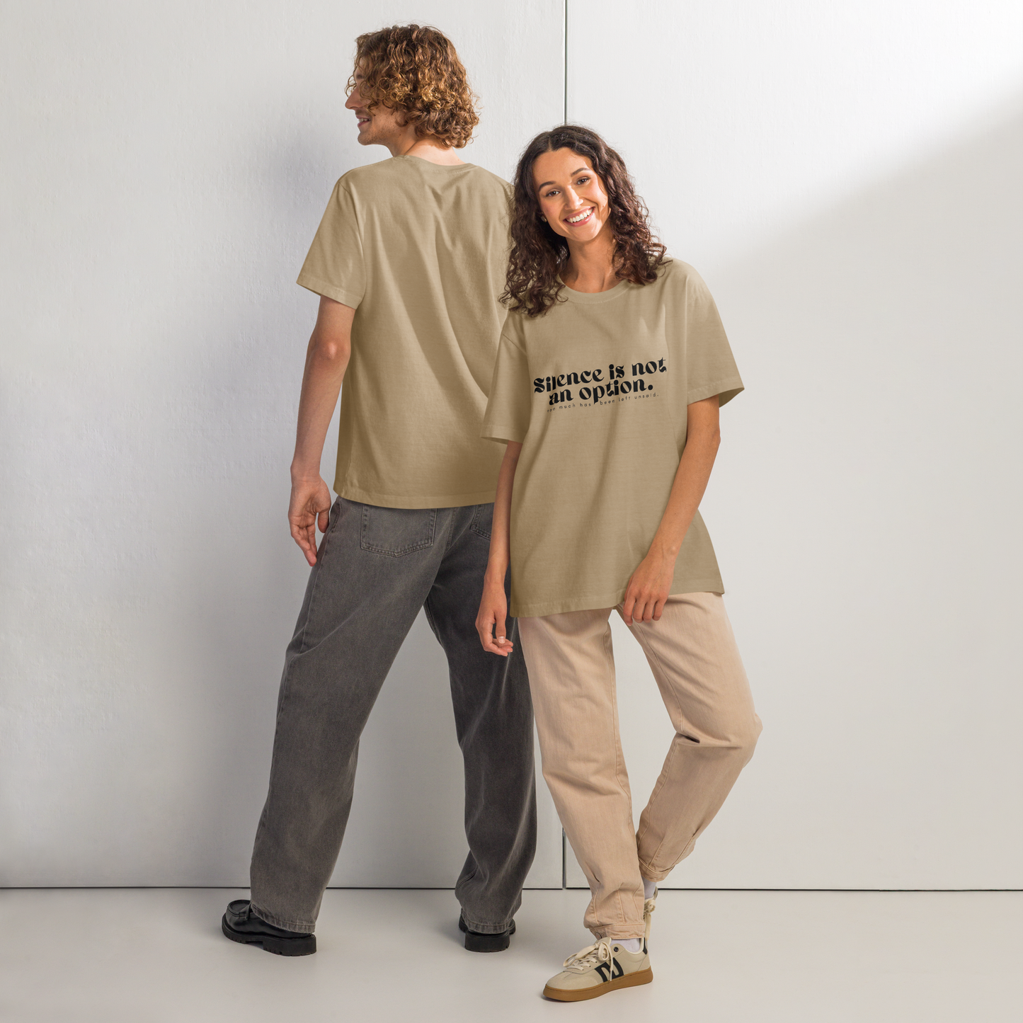 Two people wearing beige t-shirts with text, standing side by side against a white wall.