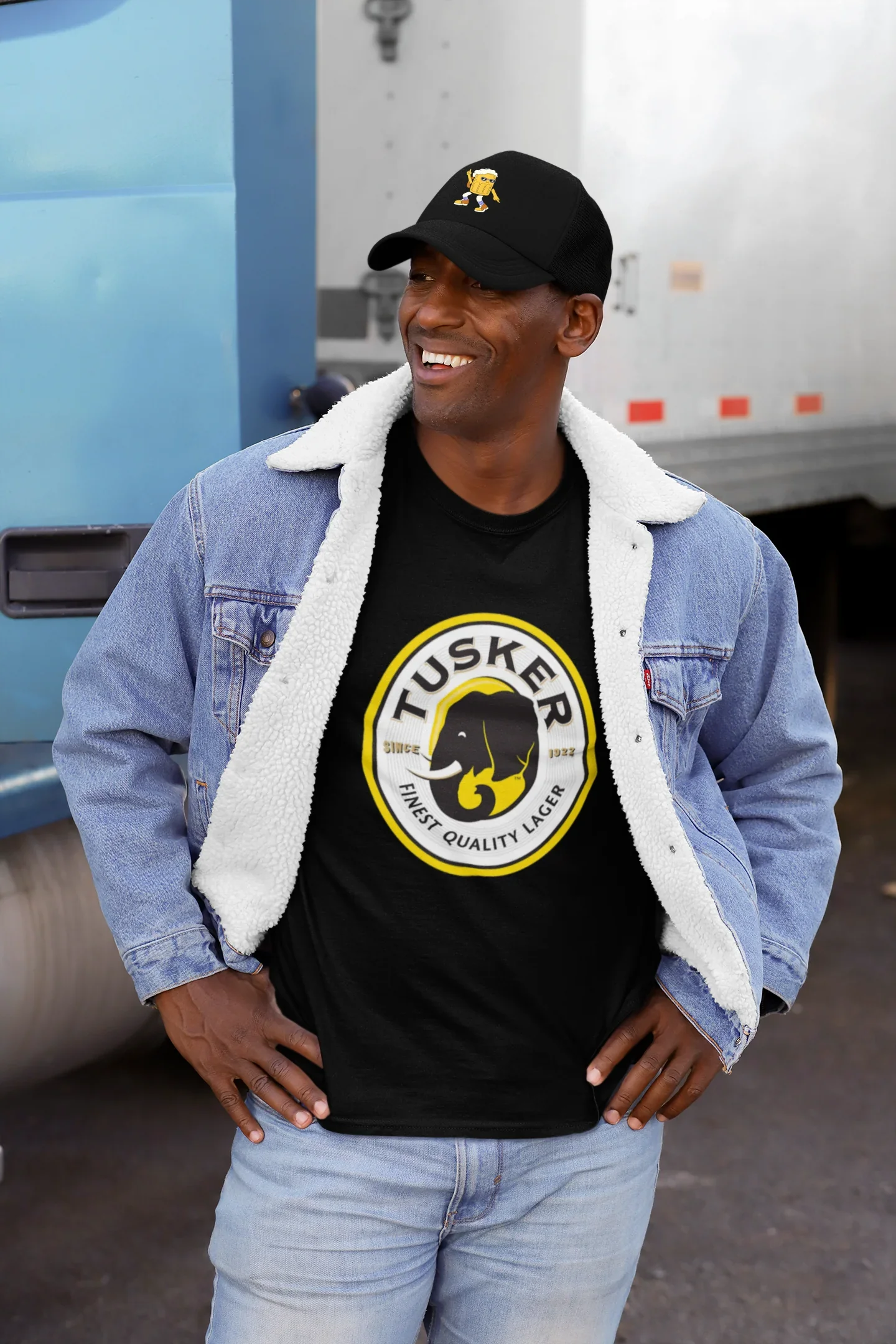 Man wearing a black Tusker t-shirt with a blue denim jacket and cap, standing in front of a truck.