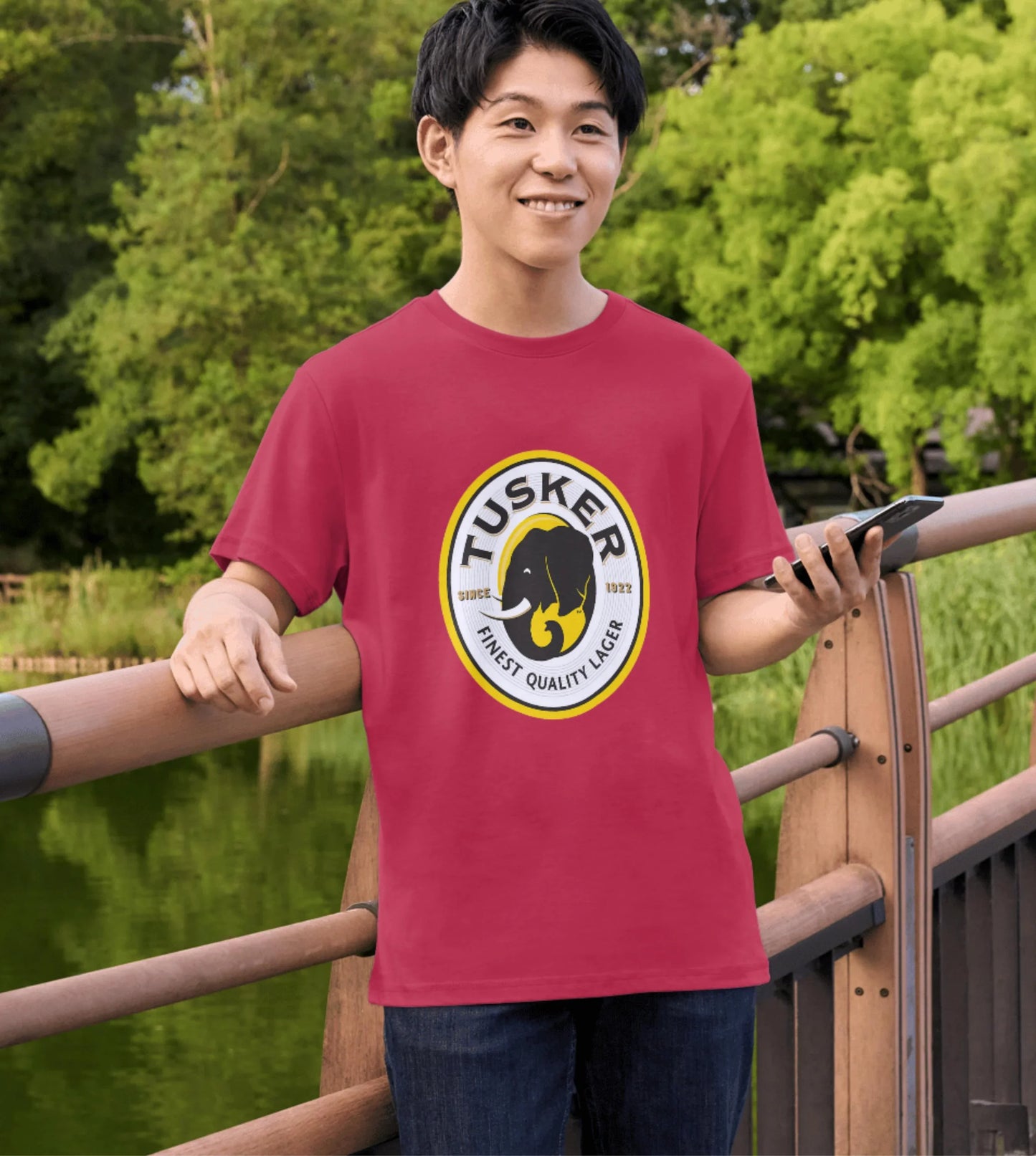 Person wearing a red Tusker t-shirt standing outdoors with greenery in the background