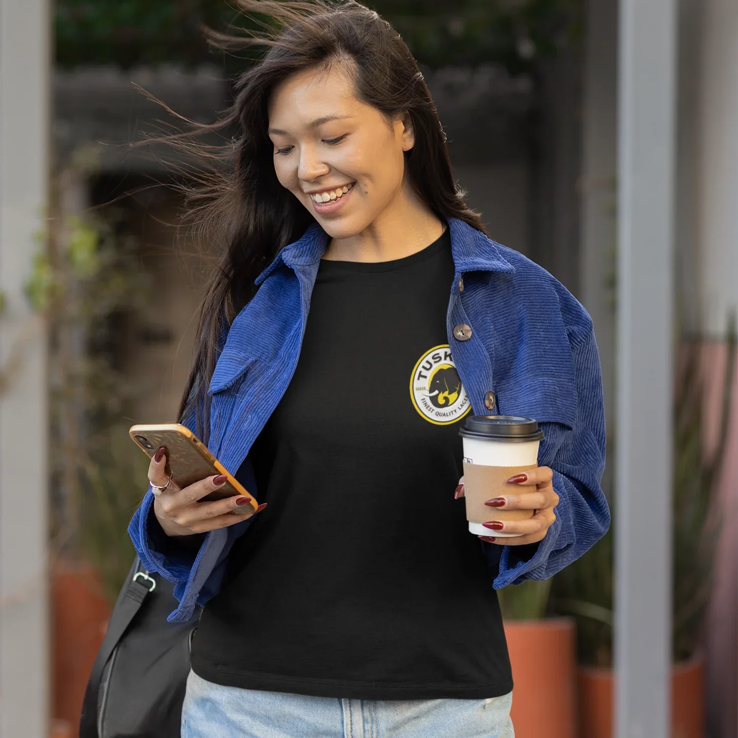 Woman holding a coffee cup and phone, wearing a black shirt with a tusker logo, outdoors.