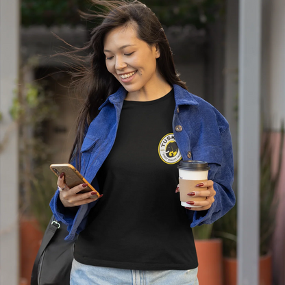 Woman holding a coffee cup and phone, wearing a black shirt with a tusker logo, outdoors.