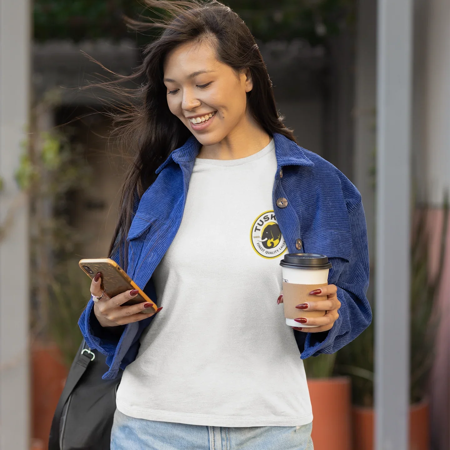 Woman holding a coffee cup and phone, wearing a white t-shirt with a logo, outdoors.
