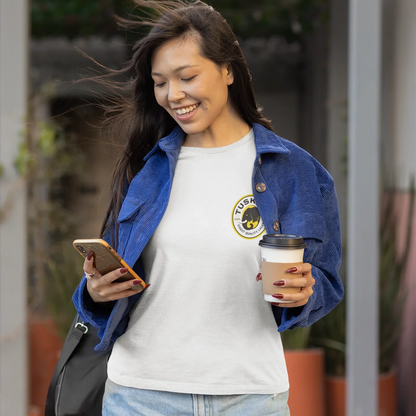 Woman holding a coffee cup and phone, wearing a white t-shirt with a logo, outdoors.