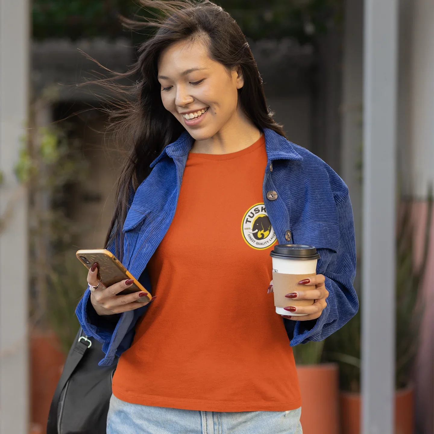 Woman holding a coffee cup and phone, wearing an orange shirt with a  tusker logo and blue jacket.