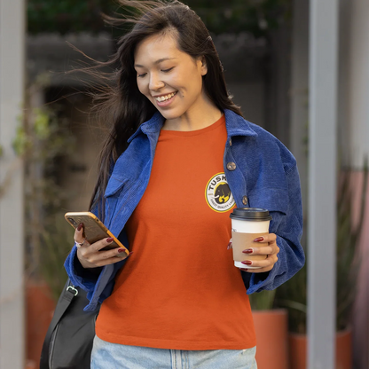 Woman holding a coffee cup and phone, wearing an orange shirt with a  tusker logo and blue jacket.