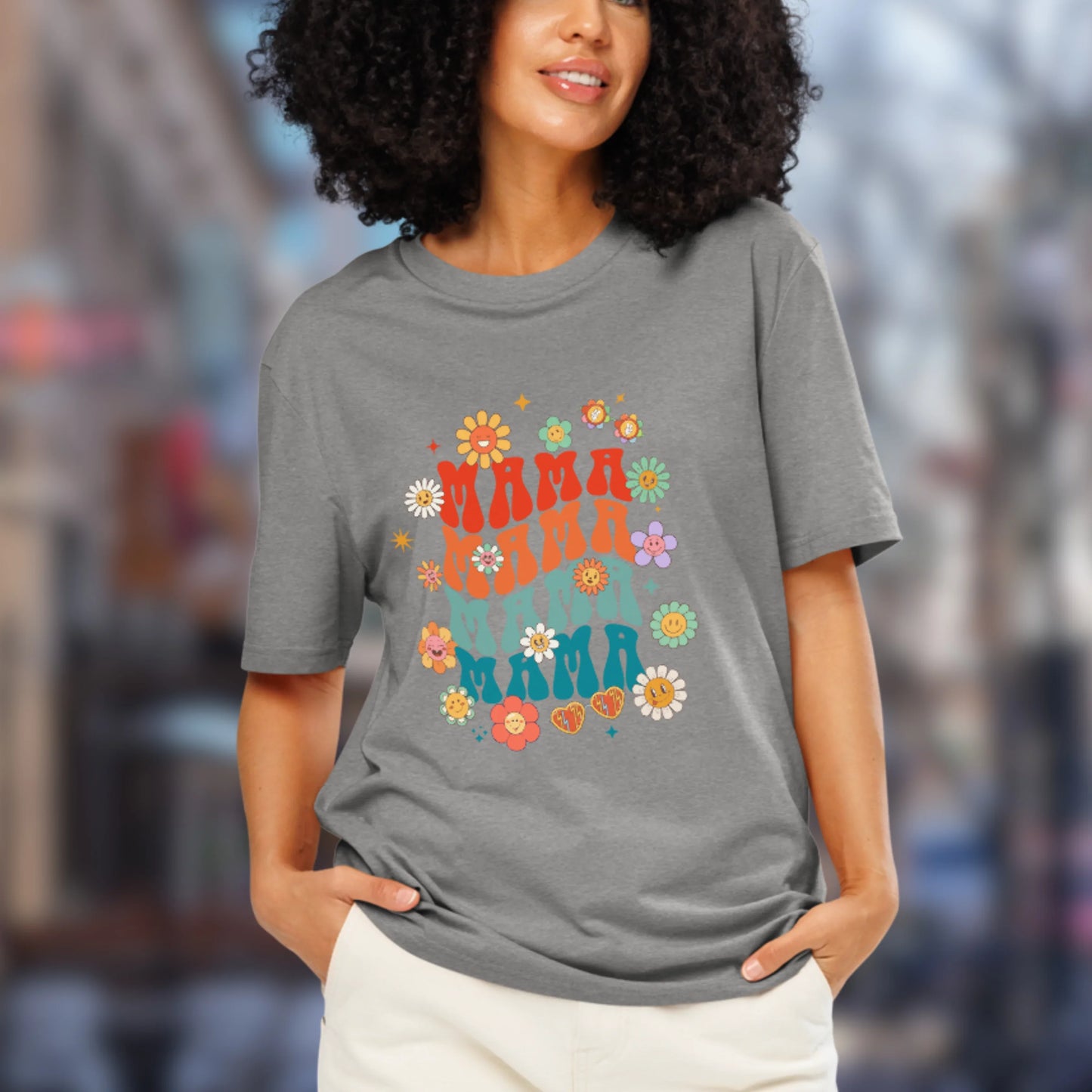 Woman wearing a gray t-shirt with colorful 'Mama' text and floral designs, standing in an urban setting.