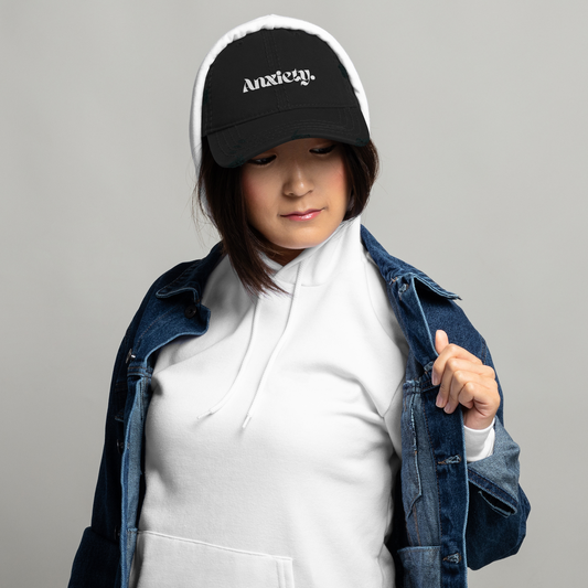 Person wearing a black cap with 'Anxiety' text and a denim jacket on a gray background