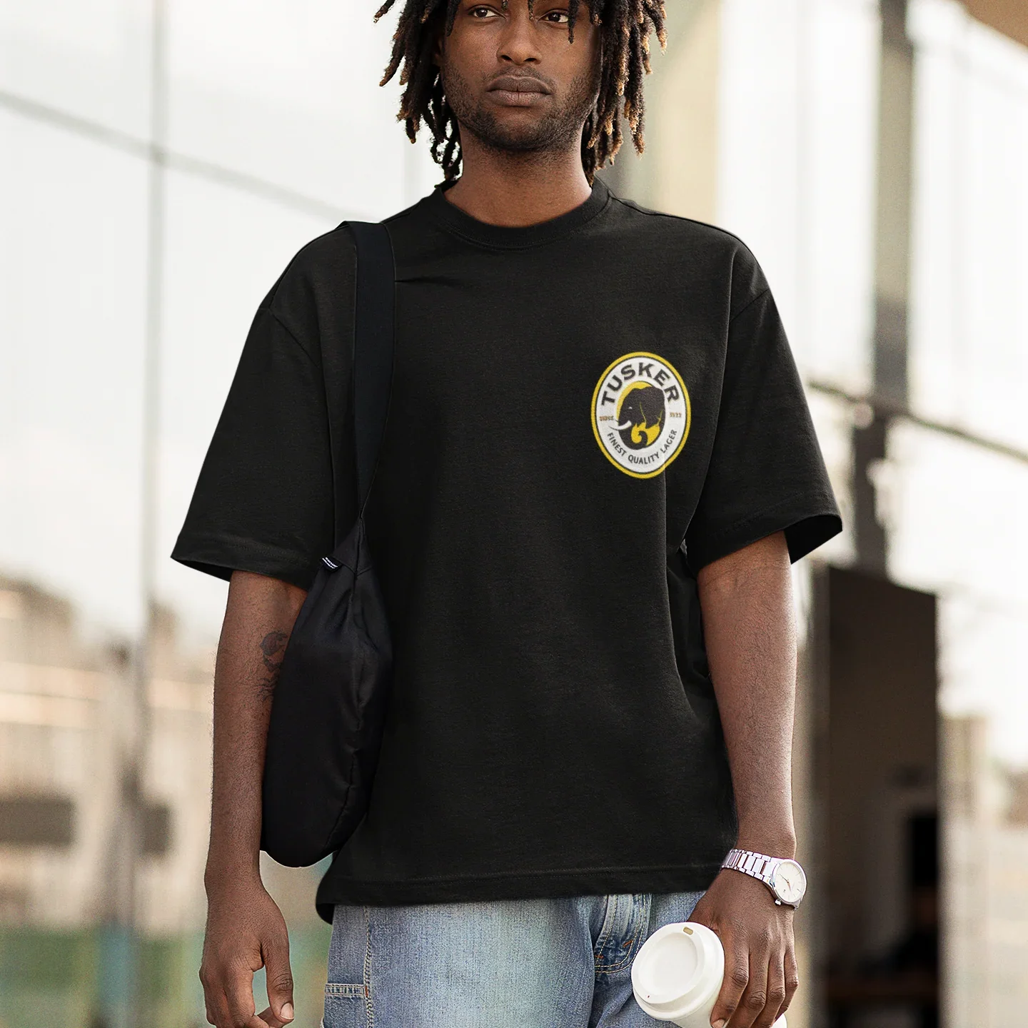 Person wearing a black t-shirt with a tusker logo, holding a bag and a cup, standing outdoors.