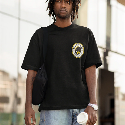 Person wearing a black t-shirt with a tusker logo, holding a bag and a cup, standing outdoors.