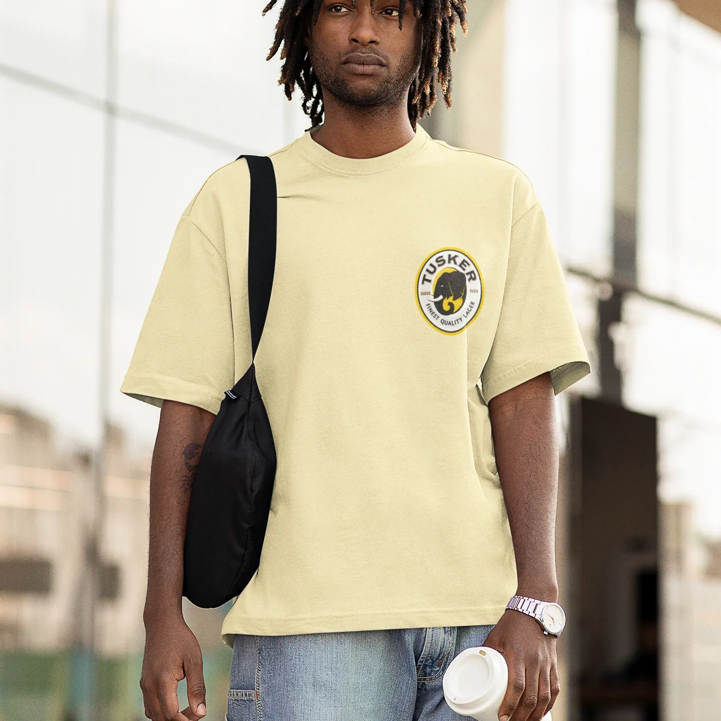 Person wearing a light yellow t-shirt with a tusker logo, carrying a black bag and holding a white cup.