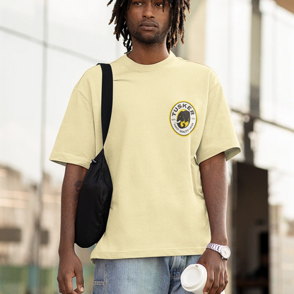 Person wearing a light yellow t-shirt with a tusker logo, carrying a black bag and holding a white cup.