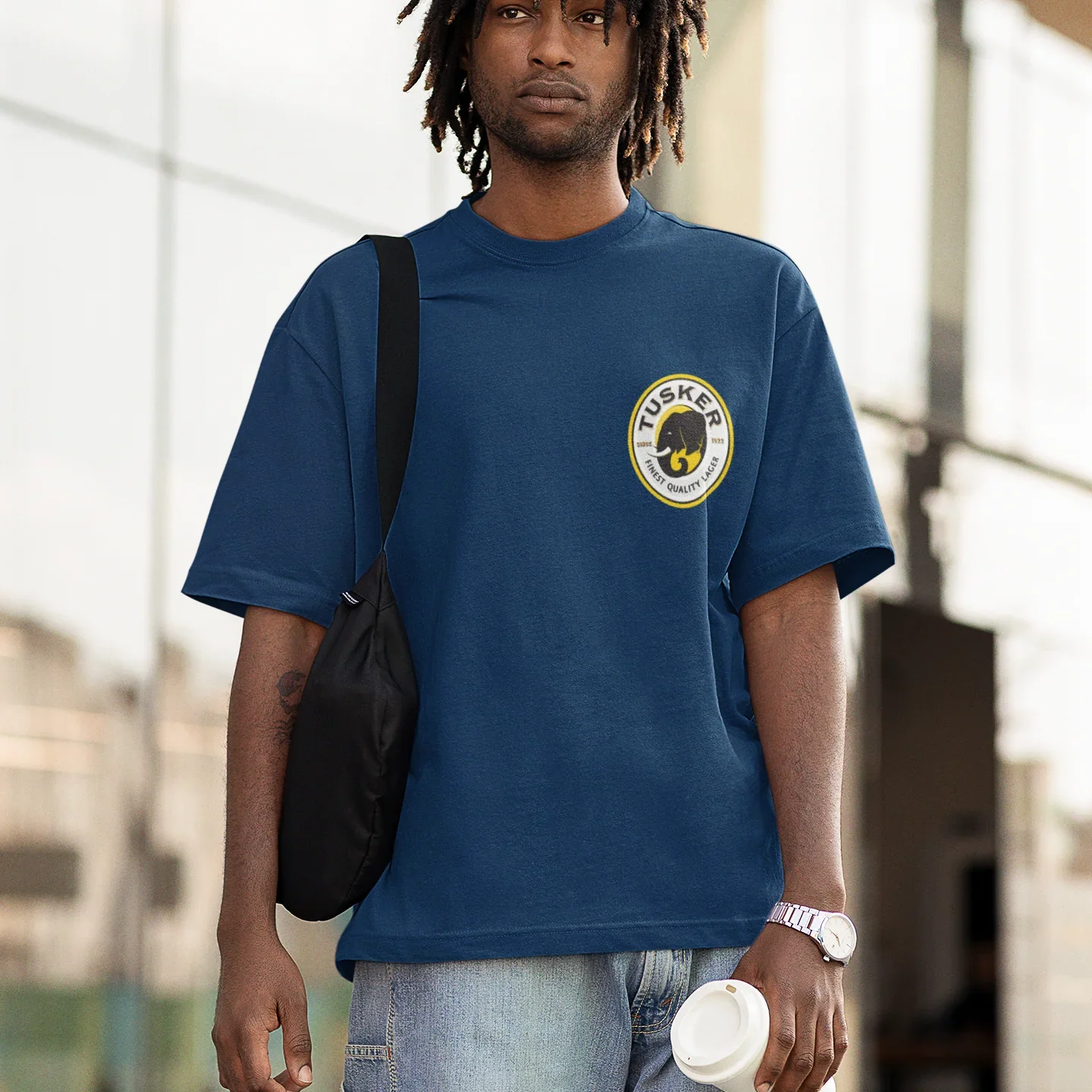 Man wearing a blue t-shirt with a logo, carrying a black bag and holding a white cup.
