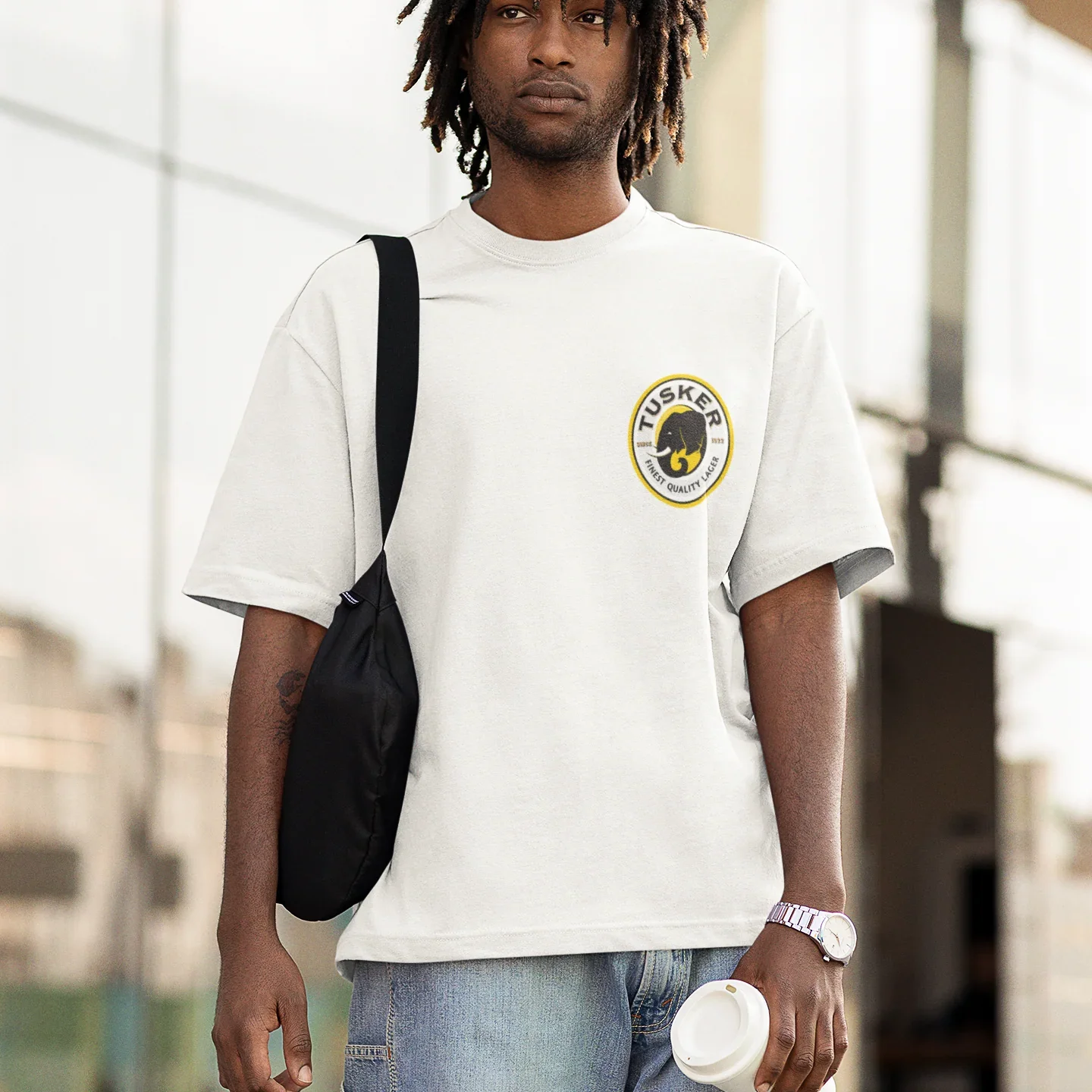 Man wearing a white t-shirt with a tusker logo, carrying a black bag and holding a cup.