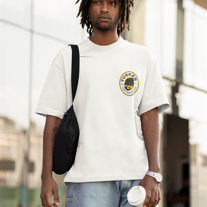 Man wearing a white t-shirt with a tusker logo, carrying a black bag and holding a cup.