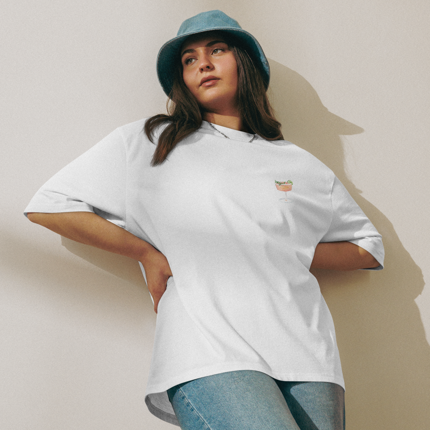 Person wearing a white t-shirt and blue jeans with a green bucket hat against a beige wall.